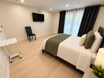 Bedroom with a bed, wall TV and soft natural light through tall curtains